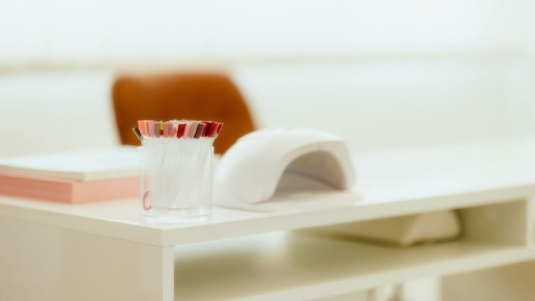 Lámpara de uñas LED blanca sobre una mesa de manicura clara, junto a un vaso con pinceles