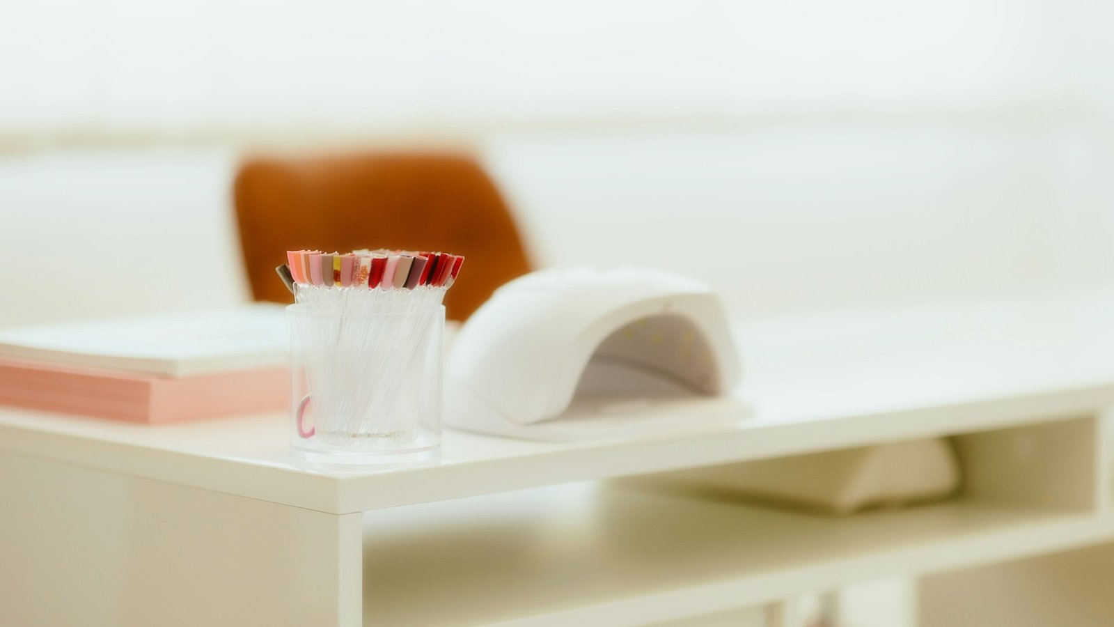 Lámpara de uñas LED blanca sobre una mesa de manicura clara, junto a un vaso con pinceles