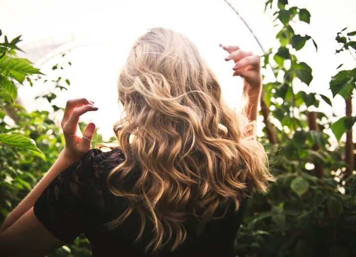 Mujer de espaldas con una melena larga rubia peinada con ondas, entre plantas verdes