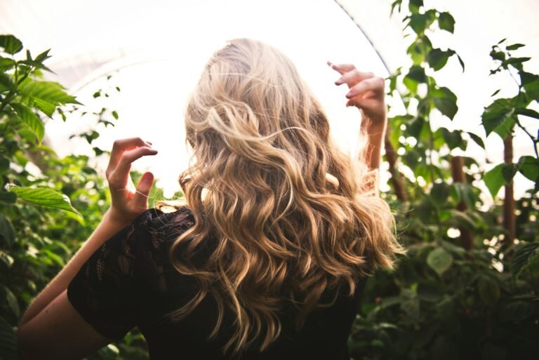 Mujer de espaldas con una melena larga rubia peinada con ondas, entre plantas verdes